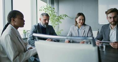 A group of people in business suits sit at a table in a modern office and communicate by videoconference with a customer using a laptop standing on the table, giving a presentation of their services video