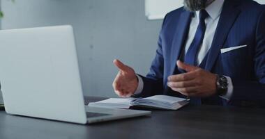 A respectable man of European appearance with a gray beard, dressed in a business suit, makes notes in a notebook while talking on a call on a laptop with a serious look, in a modern office video