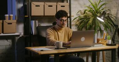 Young handsome man of European appearance with a serious look works at a laptop sitting at a table in a modern loft-style office, on the background of a rack with cardboard boxes video