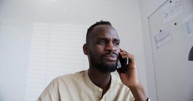 black guy with a beard in a shirt with a short afro hairstyle speaks by phone while sitting in a bright modern office video