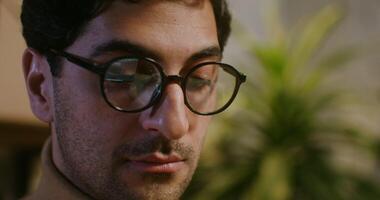 Young curly haired brunette man in glasses with stubble on his face with a serious look works at a laptop sitting at a table in the office, close-up video