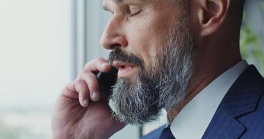 A man with a gray beard of European appearance, dressed in a business suit with a tie with a serious look, is talking on a mobile phone, close-up video