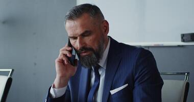A respectable man of European appearance with a gray beard, dressed in a business suit, talking on a mobile phone with a serious look, sitting at a table in the office video