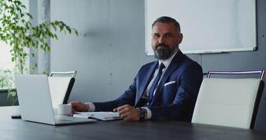 A respectable man of European appearance with a gray beard, dressed in a business suit with a serious look, looks directly at the camera, sitting in front of an open laptop in a modern office video