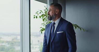 uma respeitável homem do europeu aparência com uma cinzento barba, endireita dele o negócio jaqueta, em pé perto a janela dentro uma moderno à moda escritório e encarando para dentro a distância video