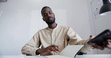 black guy with a beard in a shirt, with a short afro hairstyle seriously speaks by phone makes notes in a notebook while sitting in a bright modern office. video