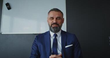 chat, front camera. A man of European appearance with a gray beard, dressed in a business suit, looks directly at the camera and smiles while talking on chat in a modern office video