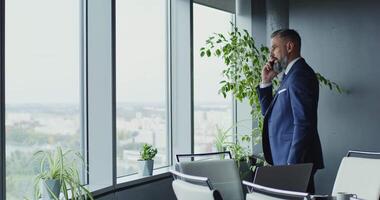 Serious middle-aged man of European appearance, dressed in a business suit, walks up to the window while talking on a mobile phone in a modern stylish office video