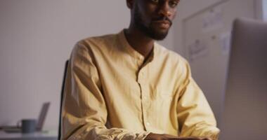 black guy with a beard in a light shirt, with a short afro hairstyle typing on a laptop and smiles in an office under under the light of a table lamp video