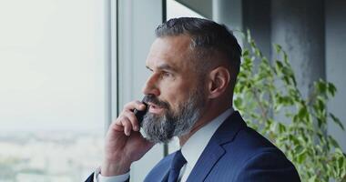 A man of European appearance, middle-aged, dressed in a business suit with a tie with a serious look, is talking on a mobile phone, standing near the window, close-up video