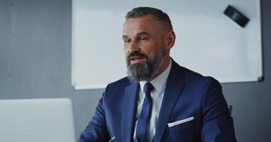 A respectable man of European appearance with a gray beard, dressed in a business suit, smiles while talking on a call on a laptop and takes notes in notebook sitting at table in the office video