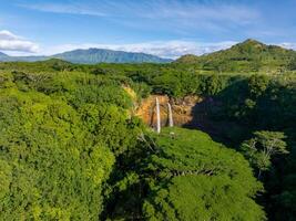 Twin streams of Wailua Falls cascade into a hidden pool, surrounded by dense tropical vegetation and rugged mountains under a clear blue sky. photo