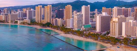 Aerial view of Waikiki Beach in Honolulu, Oahu, Hawaii, at sunset. Turquoise waters, sandy shores, palm trees, and high rise buildings are visible. photo