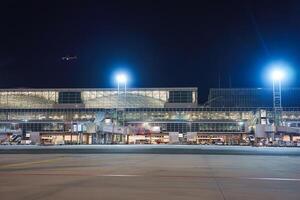 Night view of a multi level airport terminal with bright lights, jet bridges, a glass facade, a runway, and a small aircraft in the sky. photo