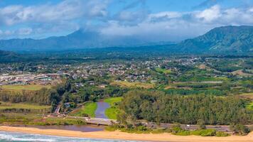 Aerial image of Kauai island, Hawaii, showing a sandy beach, winding river with a bridge, a town, verdant fields, and the Napali cliffs in the background. photo