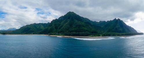 Aerial view of Kauai Island's Napali cliffs with lush greenery, sandy beach, ocean waves, rugged peaks, and a partly cloudy sky. photo