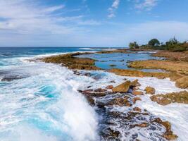 Rugged rocky shores meet vibrant blue ocean waves on Oahu island. Tide pools and sparse greenery line the coastline under a partly cloudy sky. photo