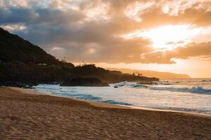 A serene Hawaiian beach at sunset with golden light, rugged cliffs, lush greenery, gentle ocean waves, and small structures in the background. photo