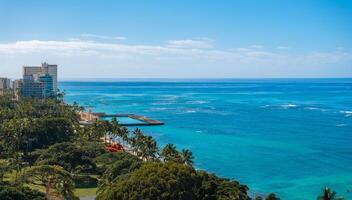 Aerial view of Waikiki Beach in Honolulu, Oahu, Hawaii, featuring turquoise waters, a small pier, high rise buildings, and lush greenery with palm trees. photo