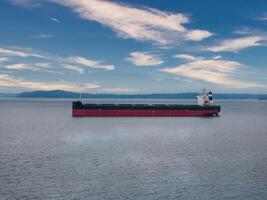 A red and black cargo ship with a white superstructure floats in a calm bay. Low hills line the distant shoreline under a partly cloudy sky. photo