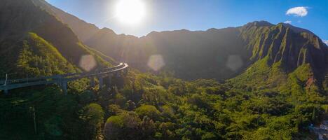 Aerial view of a green valley on Oahu island, Hawaii, with jagged mountain ridges, a curving elevated highway, and sunlight casting a warm glow. photo