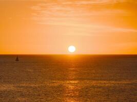 A tranquil sunset over the ocean in Oahu, Hawaii, with a small sailboat on the left. The sky glows in warm orange and yellow hues with scattered clouds. photo