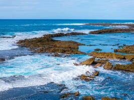 Waves crash against rugged rocks on Oahu's North Shore, creating white foam. Calm pools and distant figures enjoying the water add scale and activity. photo