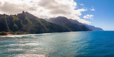 Aerial view of the Napali Coast on Kauai Island, Hawaii, with jagged green cliffs, turquoise waters, gentle waves, and partly cloudy skies. photo