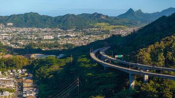 A highway curves through lush green hills on Oahu island, Hawaii, with a sprawling urban area and a triangular mountain peak in the background. photo