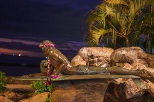 A bronze statue of a surfer on a surfboard adorned with flower leis, set against rocks, tropical plants, and an ocean backdrop under a darkening sky. photo