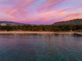 Aerial view of Waikiki Beach in Honolulu, Oahu, Hawaii, at sunset. Palm trees, clear waters, cityscape, and Diamond Head crater are visible. photo