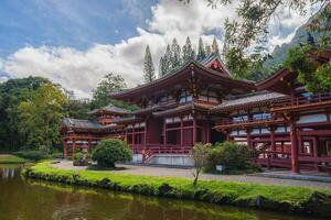 The Byodo In Temple in Oahu, Hawaii, surrounded by greenery, a serene pond, and rugged mountains under a partly cloudy sky. Traditional Japanese design. photo