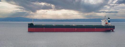 A large cargo ship with a red hull floats on calm waters near a tree lined shoreline. Sunlight breaks through scattered clouds, creating a serene scene. photo