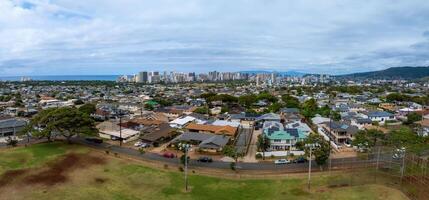 Aerial view of a residential area in Honolulu, Hawaii, with homes, a park, and greenery. The skyline and Pacific Ocean are visible, framed by mountains. photo