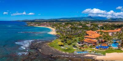 Aerial image of a Kauai island resort with orange tiled roofs, a pool, palm trees, a sandy beach, turquoise ocean, and distant green mountains. photo