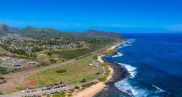 Aerial view of Oahu, Hawaii, featuring a blue ocean, rugged shoreline, winding road, green fields, scattered buildings, and rolling hills in the background. photo