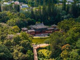The Byodo In Temple in Oahu, Hawaii, features intricate architecture with red accents, set amidst a manicured garden, pond, and tall trees. photo