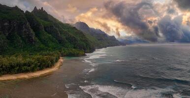 Aerial view of Kauai's Napali Coast with jagged green cliffs, a sandy beach, turquoise waters, lush vegetation, and a vibrant sky with dramatic clouds. photo