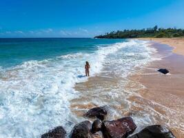 A tranquil beach on Kauai island features golden sand, turquoise waves, lush greenery, dark rocks in the foreground, and a person in shallow water. photo