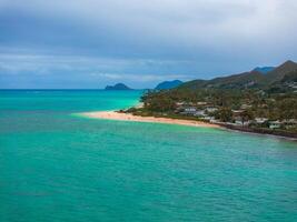 Turquoise ocean meets sandy beach lined with palm trees and homes, with lush hills and a small offshore island under a cloudy sky in Oahu, Hawaii. photo