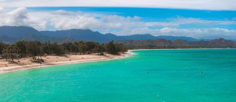 Aerial view of a beach on Oahu island, Hawaii, featuring turquoise waters, sandy shoreline, scattered trees, and lush green mountains under a cloudy sky. photo