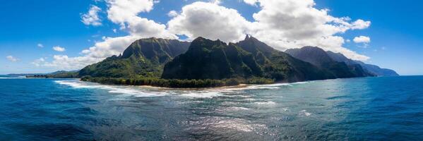 Aerial view of Kauai's Napali Coast with jagged green cliffs, sandy beach, turquoise ocean waves, and a partly cloudy sky creating vivid contrasts. photo