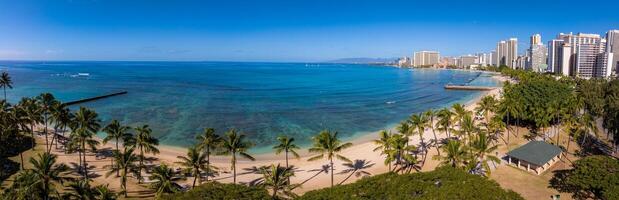 Aerial view of Waikiki Beach in Honolulu, Oahu, Hawaii, featuring turquoise waters, sandy shores, palm trees, piers, and high rise buildings under a clear sky. photo