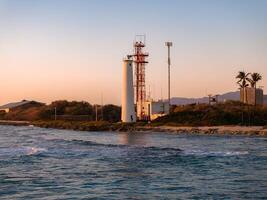 A white lighthouse with red accents near a rocky shoreline at sunset, surrounded by sparse vegetation, palm trees, and distant mountains on Oahu. photo