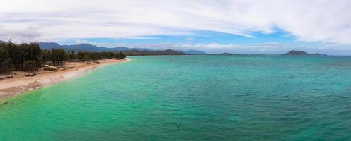 Aerial view of a beach on Oahu island, Hawaii, with turquoise waters, sandy shoreline, tree lined border, distant mountains, and an island on the horizon. photo
