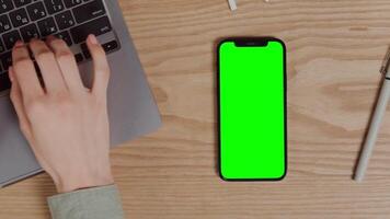 Close-up of a mobile phone with a green screen lying on a table next to a woman's hand typing on a laptop. Chroma key, mockup, copy space. in 4k format, red komodo. video