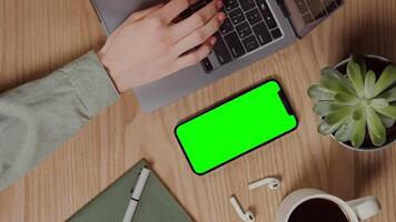 Close-up of a smartphone with a green screen lying on a table next to female hands typing on a laptop, vertical . Chroma key, mockup, copy space. in 4k format, red komodo. video
