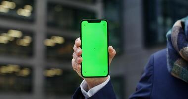 A warmly dressed man holds a green screen phone as he stands outside an office building. Copy space, mockup, close-up of the phone, no face. in 4k, red komodo video