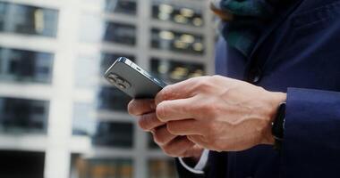 Close-up of male hands with smartphone. A businessman is typing on a mobile phone while standing outdoors near an office building. Unrecognizable person. in 4k, red komodo video