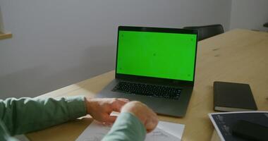 A young man of European appearance is working on a drawing, sitting in front of an open laptop with a green screen, copy space, chroma key. Young architect's desk with many papers and blueprints video